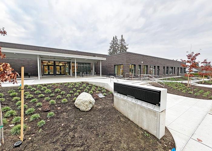 a long one-story building includes a breezeway in front of doors and a dark brick section. a walkway leads between newly planted landscaping toward the entrance with an electronic sign beside it